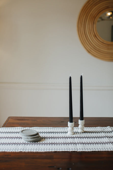 A pair of black candlesticks styled on a dining table with modern, white stone candle holders, a set of four concrete coasters, and a geometric black and white table runner